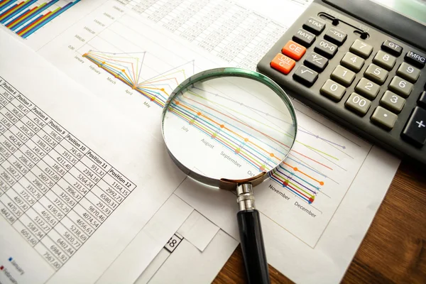 Business accessories (notebook, calculator, planchette, tablet, fountain pen, glasses) and graphics, tables, charts on white sheets on office desk. Soft focus.