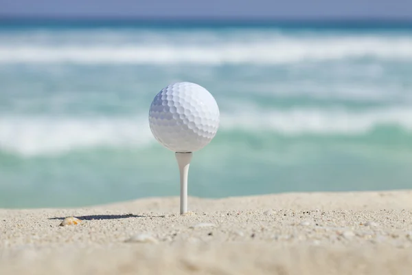Golf ball on tee in sand beach with ocean waves behind