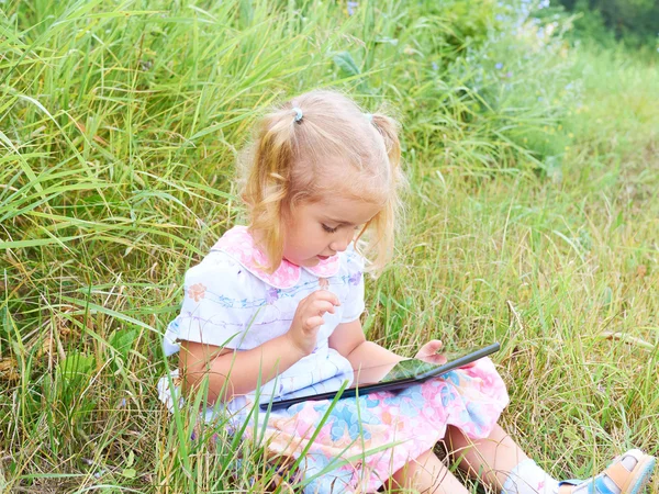 Little girl sitting in park on sunny day and play with tablet pc