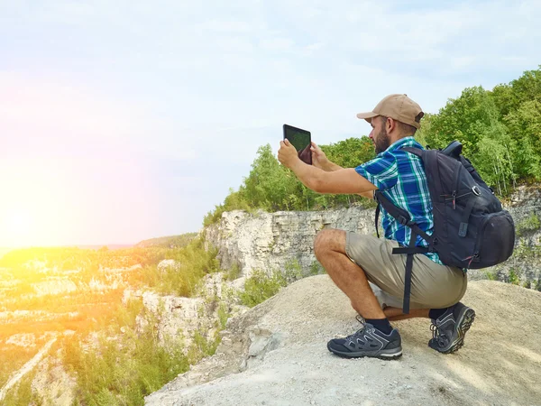 Man tourist uses tablet computer sitting on the edge of a cliff