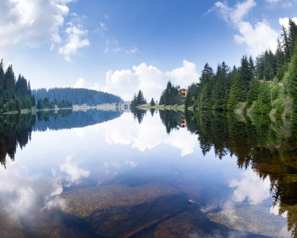Blue expanse of clear forest lake with huge stones on the bottom, pine forest and cloudy sky and their reflection in the water early in the morning in the Rhodope Mountains