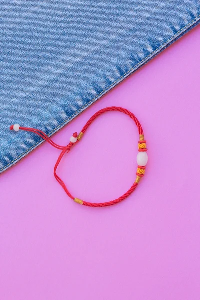 Bracelet red thread and blue jeans on a pink background. fashionable jeans top view