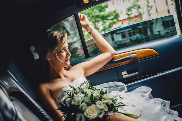 Portrait of a beautiful blonde bride sitting in  wedding car and looking at somebody or something through the window. Wedding day. Daylight