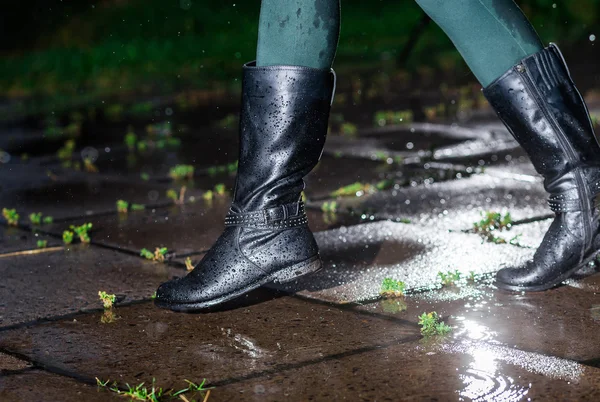 Female legs in black leather high boots on cobble  the edge of rain puddle, closeup. Concept  protection against rainy weather, waterproof footwear