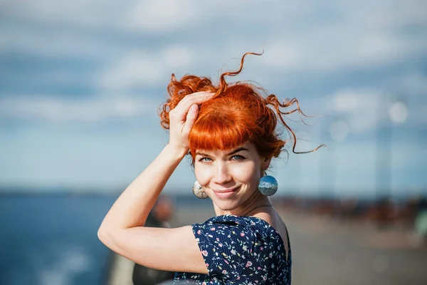 Beautiful young redhead woman smiling and holding her hands in her hair with hear eyes closed on the background of embankment