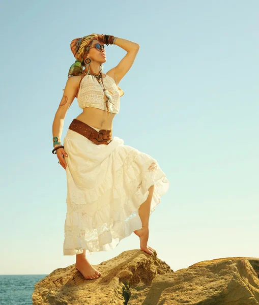 Fashion shot of a beautiful boho style woman standing on a rock near sea. Boho outfit, hippie, indie style