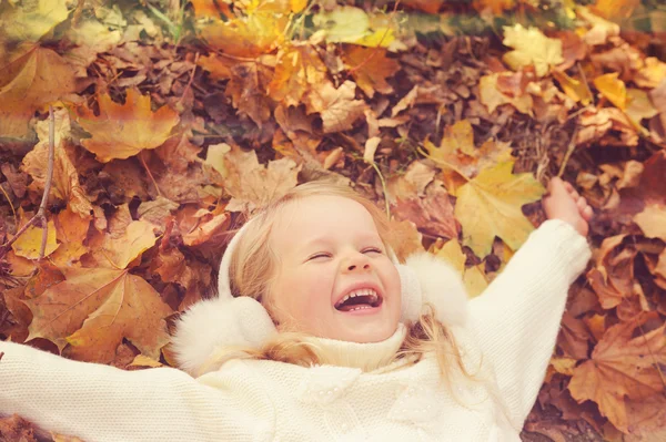 Little blonde girl portrait lying on autumn yellow maple leaves outstretched hands and smiling.