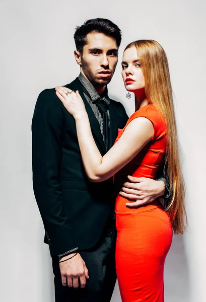Young fashion man and woman against white wall, looking at the camera. She hugs him. Indoor. Warm color.