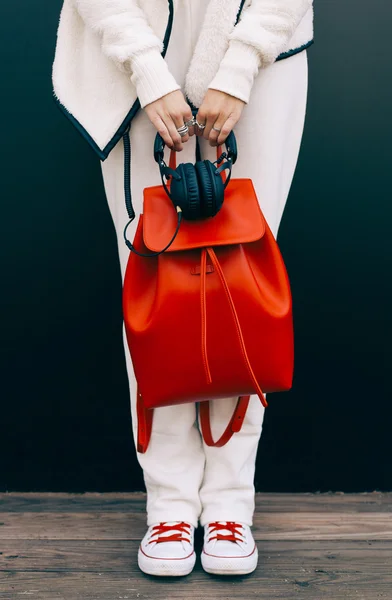 Fashionable beautiful big red backpack and headphones on the arm of the girl in a fashionable white sports suit and Trainers, posing near the wall on a warm summer night. Warm color. Close up