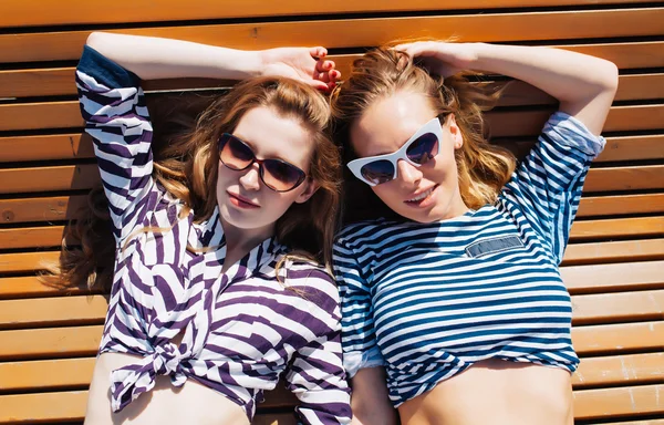Close up lifestyle summer portrait of two girls friends relaxed and getting sunbathe, laying on the beach, wearing bright marine costumes and stylish sunglasses. Yacht. Sea, summer.