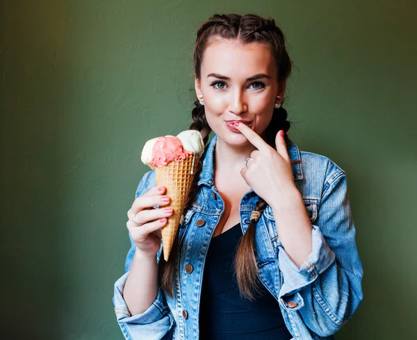 Beautiful girl with braids sitting in a cafe and eating huge multi-colored ice cream in a waffle cone. Licking finger