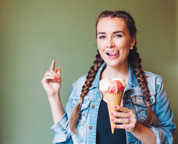 Beautiful girl with braids sitting in a cafe and eating huge multi-colored ice cream in a waffle cone. Smiling at the camera and licking lips.