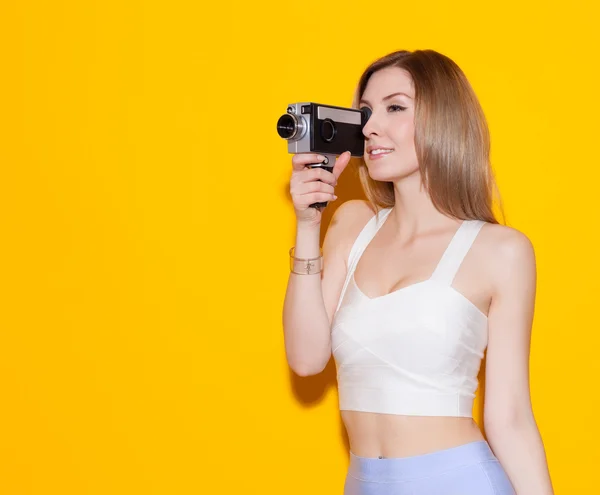 Beautiful fashionable girl posing and holding a vintage movie camera in a top and a skirt on a yellow background in the studio. Gorgeous Woman Portrait. Stylish Haircut and Makeup. Hairstyle. Make up.