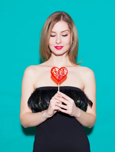 Fashionable beautiful girl holding a red candy heart and look at his. In a black dress on a green background in the studio. Fashion Beauty Girl. Gorgeous Woman Portrait. Stylish Haircut and Makeup. Ha