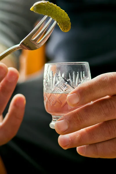 Glass of vodka and pickled cucumber on a fork in male hands.