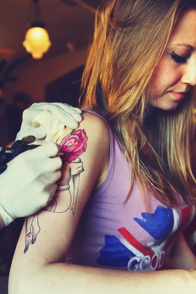 Tattooer showing process of making a tattoo on young beautiful hipster woman with blonde hair hand. Tattoo design in the form pin-up girl with flower head