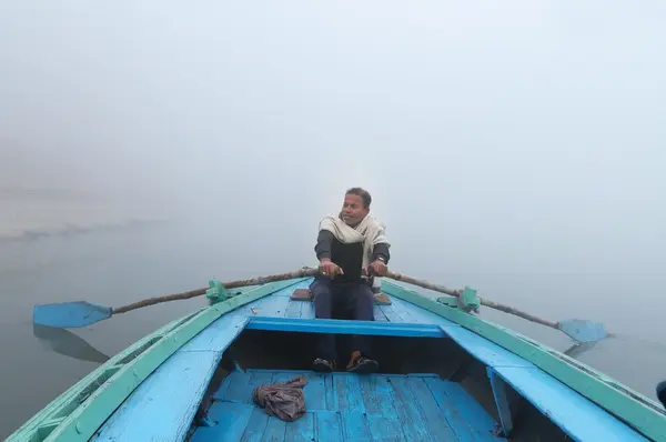 Indian man sailing on the boat on sacred river Ganges at cold foggy winter morning