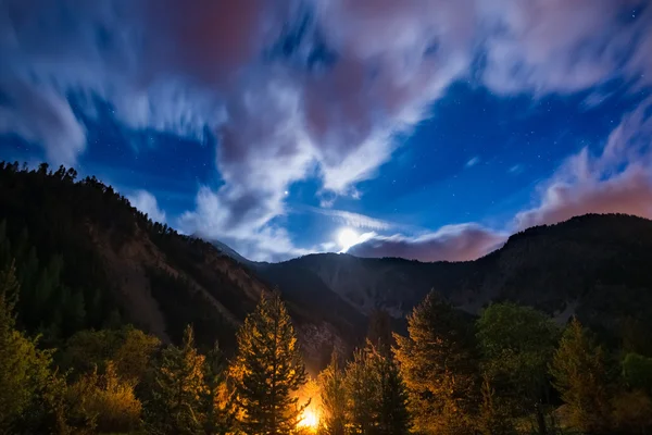The starry sky with blurred motion clouds and bright moonlight, captured from larch tree woodland, glowing by burning fire. Expansive night landscape in the European Alps. Concept of adventure into th