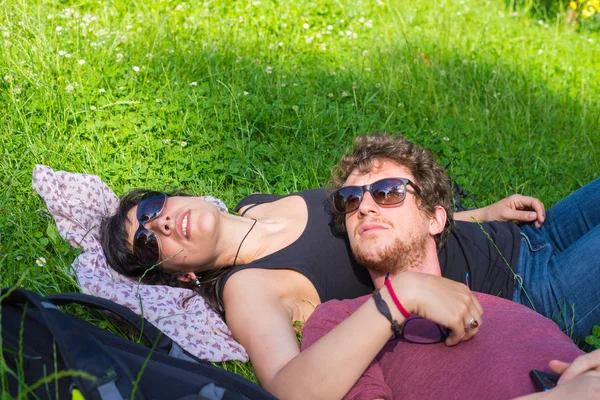 Young adult couple lying down on lush green grass in park, relaxing, embracing. Real people, natural color. Conpept of happy relationship and summer lifestyle.