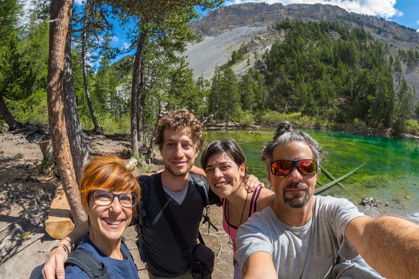 Four young people taking selfie in idyllic landscape with green lake, conifer woodland and mountains in background. Scenic fisheye distortion. Concept of traveling people and nature beauty exploration.