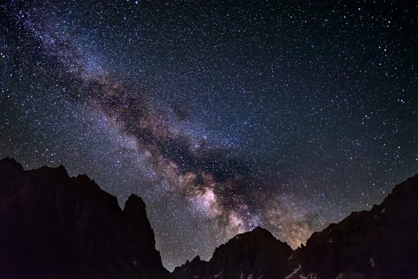 The colorful glowing core of the Milky Way and the starry sky captured at high altitude in summertime on the Alps. Scenic snowcapped mountain silhouette.