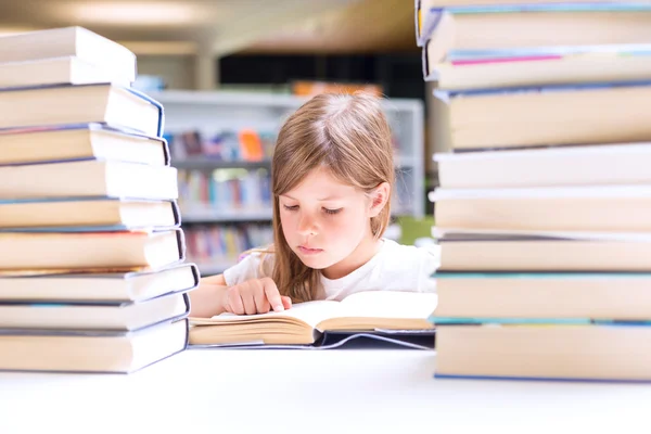 Little girl reads a book surrounded with piles of books in libra