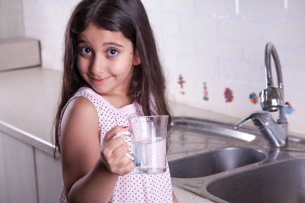 One beautiful middle eastern little girl with pink dress and long dark brown hair and eyes on white kitchen,helping parents to wash dishes and drinking water and smiling looking at camera.