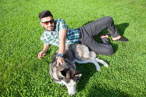 One fashion middle eastern man with beard and fashion hair style is resting and enjoy on beautiful green grass day time and playing with his with Hasky dog. looking at camera with smile and happiness.