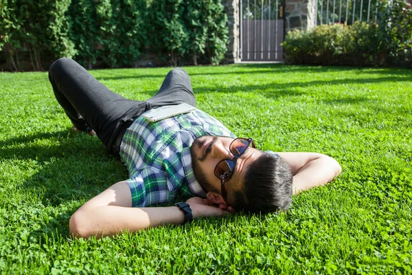 One fashion middle eastern man with beard, fashion hair style is resting on beautiful green grass day time. young arab businessman 20-30 years, resting after hard work. smart phone, tablet.
