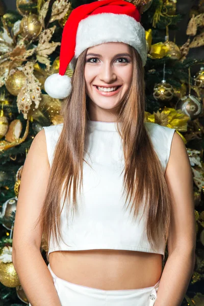 Young funny beautiful fashion model with dark eyes, brown hair and santa hat celebrating new year at home. new year decoration, with positive emotion posing and  looking at camera. yellow gold decor