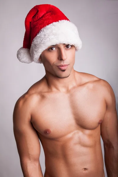 Beautiful sexy naked fashion model with new year hat and sexy muscular body, looking at camera with passion and smile. studio shot on gray background. sexy santa claus. new year sex symbol.