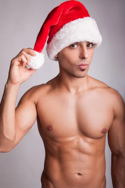Beautiful sexy naked fashion model with new year hat and sexy muscular body, looking at camera with passion and smile. studio shot on gray background. sexy santa claus. new year sex symbol.