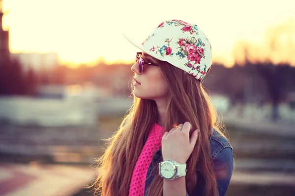 Outdoor fashion portrait of stylish swag girl, wearing swag cap, trendy sunglasses and denim jacket, amazing view of the beach at sunset.