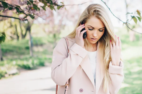 Young beautiful woman in a pink coat posing with flowers in  garden.Speak on mobile phone