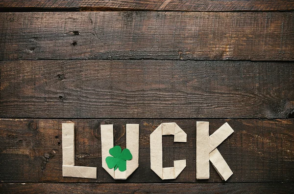 Craft paper origami LUCK lettering on dark barn wood rusctic background. Green clover leaf. St. Patrick\'s Day greeting postcard template. Space for copy, text, lettering.
