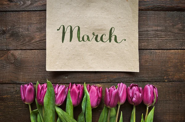 March brush nib lettering calligraphy. Pink, tulips bunch on dark barn wood planks background. Postcard template.