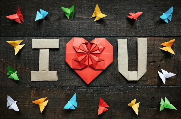 I LOVE YOU, U folded letters and red heart. Colored origami paper butterflies frame on dark brown aged wood background. Valentines day horizontal postcard template. Space for copy, text, lettering.