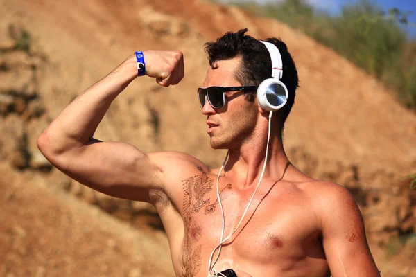 Muscle sexy wet naked young man in sunglasses on the background of rocks with headphones and music player. Shows great biceps