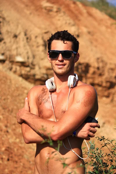 Serious naked muscular young man standing in the desert smiling, embracing himself. Wearing sunglasses and white headphones