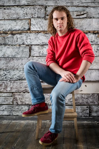 A young man in a red sweater and jeans sitting on a wooden stool