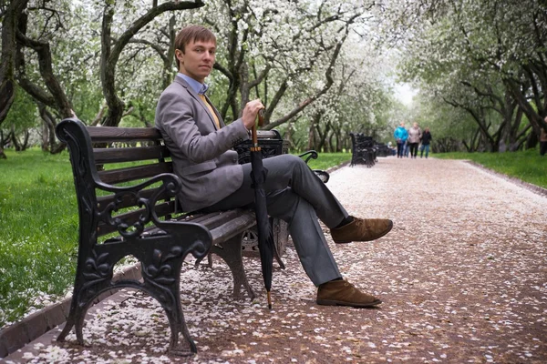 A young man in a gray suit with a black umbrella-cane on the bench in the lush apple orchard