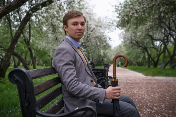 A young man in a gray suit with a black umbrella-cane on the bench in the lush apple orchard