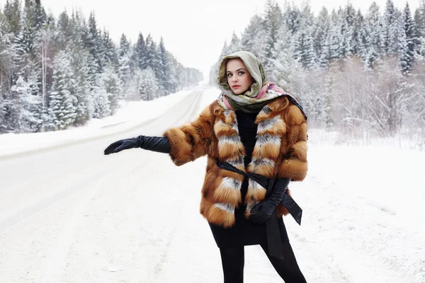 Beautiful girl in a fur coat waiting for the car on a winter road in the forest.young Woman in fashionable Fur Coat,leather gloves and scarf