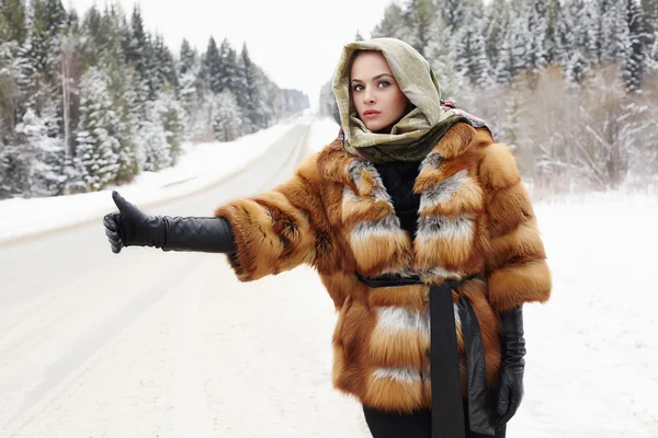 Beautiful girl in a fur coat waiting for the car on a winter road in the forest.young Woman in fashionable Fur Coat,leather gloves and scarf