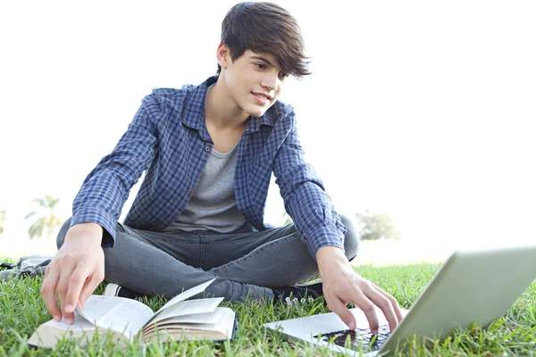 Boy in a park reading an book and using a laptop