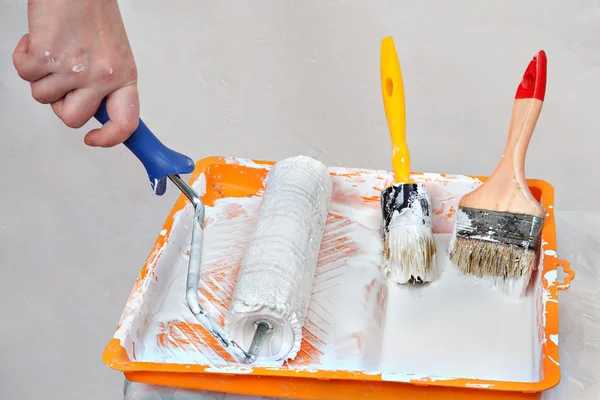 Hands painter dunks roller brush in tray of white paint.