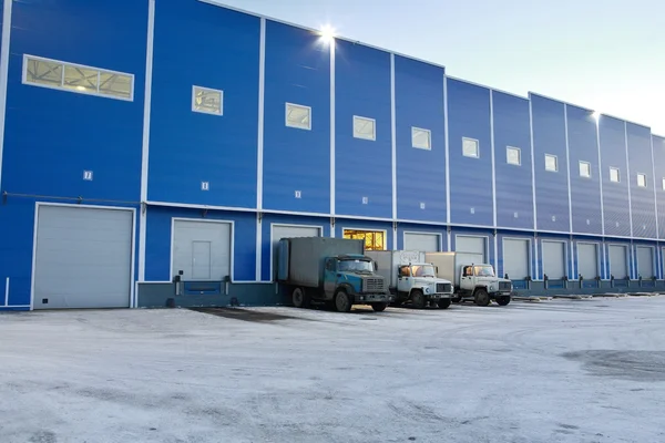 Modern Warehouse outside,  trucks are unloaded at loading docks, evening.