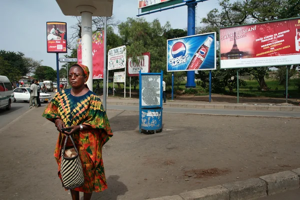 Dark-skinned Tanzanian woman stands on street surrounded by outd