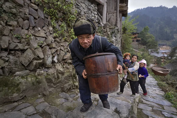 Chinese older man climbs stone mountain road with wooden barrel