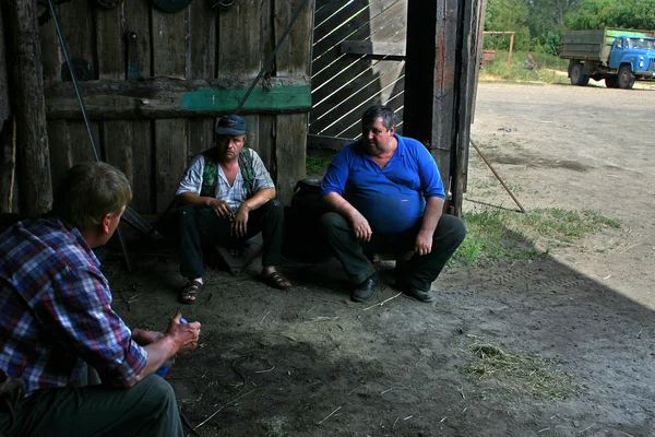 Three men, farm workers talking in shade of wooden barn.